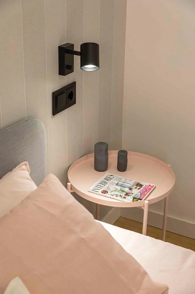  Mesita de noche redonda de color rosa pastel y aplique de pared negro de diseño en un proyecto de home staging en A Coruña.